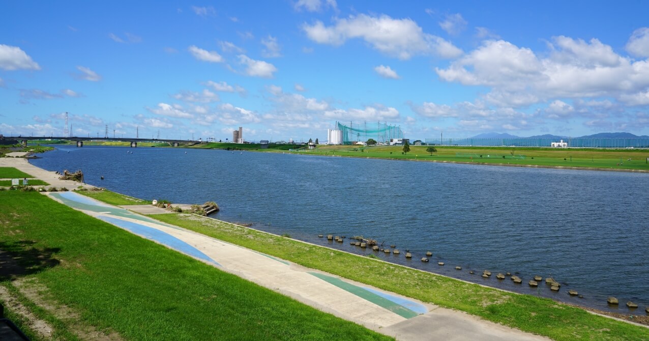 青い空と雲の下、広い川が流れ、手前には緑の土手と散歩道が広がる風景